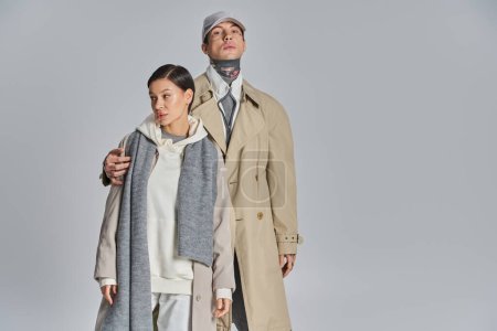 Foto de Una pareja joven y elegante con abrigos de trinchera de pie uno al lado del otro en un estudio sobre un fondo gris. - Imagen libre de derechos
