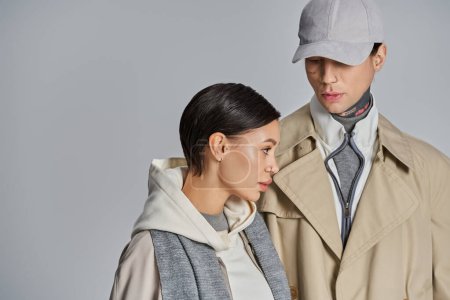 Foto de Una pareja joven y elegante en abrigos de trinchera posando con confianza en un estudio sobre un fondo gris. - Imagen libre de derechos