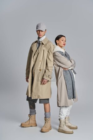 Ein stylisches junges Paar steht nebeneinander in Trenchcoats und posiert in einem Studio vor grauem Hintergrund.