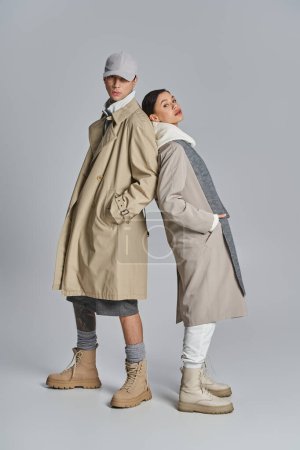 Un jeune couple élégant se tient ensemble dans des trench-coats sur fond de studio gris.