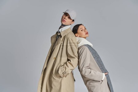 Foto de Un joven hombre y una mujer de pie lado a lado en abrigos de trinchera, exudando estilo urbano y elegancia contra un fondo gris estudio. - Imagen libre de derechos