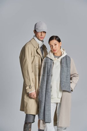 Photo for A young stylish couple stands side by side in trench coats in a studio against a grey background. - Royalty Free Image