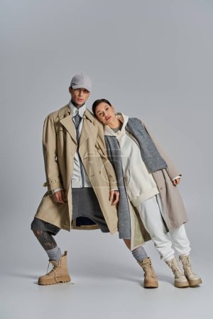 Foto de Una pareja joven y elegante está de pie lado a lado en abrigos de trinchera en un entorno de estudio sobre un fondo gris. - Imagen libre de derechos