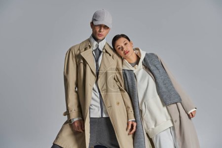 Foto de Una pareja joven y elegante se encuentra con confianza lado a lado en abrigos de trinchera contra un fondo gris. - Imagen libre de derechos