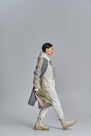 Eine junge, stylische Frau läuft selbstbewusst im Trenchcoat in einem Studio mit grauem Hintergrund.