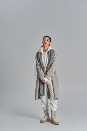 Téléchargez les photos : Une jeune femme élégante se tient avec confiance dans un trench coat sur un fond gris, respirant élégance et assurance. - en image libre de droit