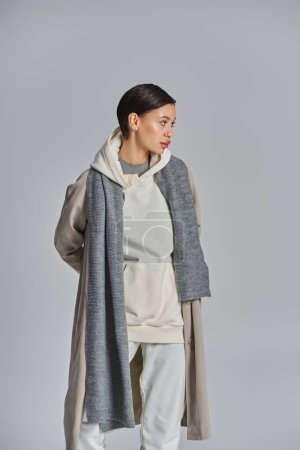 Téléchargez les photos : Une jeune femme élégante se tient avec confiance dans un trench coat sur un fond gris uni. - en image libre de droit