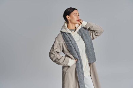 Téléchargez les photos : A young stylish woman stands in a studio, dressed in a long coat and scarf, exuding elegance and sophistication. - en image libre de droit