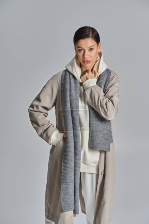 Une jeune femme élégante respire l'élégance dans un trench coat gris et un pantalon blanc sur fond de studio neutre.