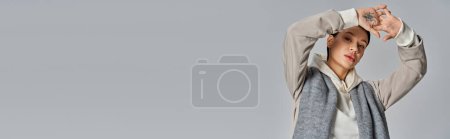 Téléchargez les photos : Une jeune femme élégante dans un trench-coat mettant en valeur une écharpe élégante enveloppée autour de son cou dans un studio sur un fond gris. - en image libre de droit