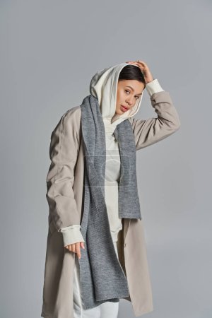 Eine junge stilvolle Frau steht anmutig in grauem Trenchcoat und weißer Hose in einem Studio mit grauem Hintergrund.