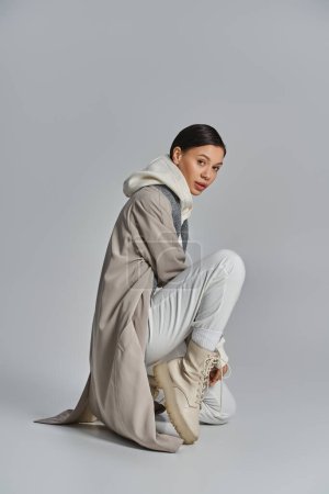 Téléchargez les photos : Une jeune femme élégante assise sur le sol, à la mode dans un trench coat, sur un fond de studio gris. - en image libre de droit
