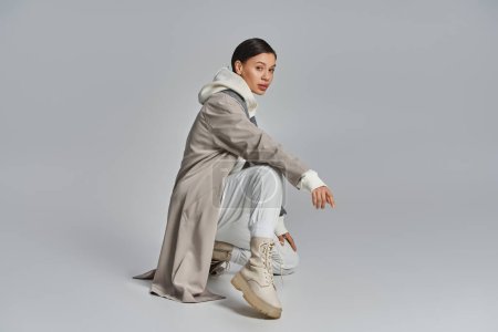 Photo for A young stylish woman in a trench coat sitting gracefully in a studio against a grey background. - Royalty Free Image
