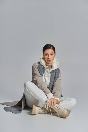 A chic young woman in a trench coat sits cross-legged on the ground, exuding a sense of calm and contemplation.
