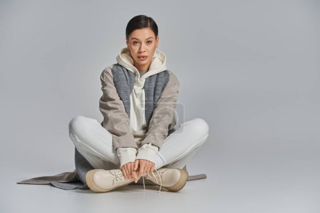 Photo for A young stylish woman in a trench coat sits peacefully on the floor, legs crossed, lost in thought in a studio setting with a grey background. - Royalty Free Image