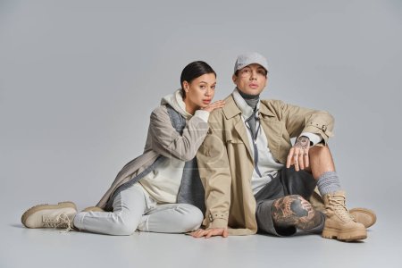 A man and a young stylish woman in a trench coat sit together on the ground, sharing a moment of connection and intimacy.