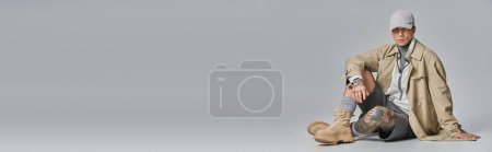 Photo for A young, tattooed man sitting on the ground, wearing trench coat, exuding a sense of thoughtfulness on a grey studio background. - Royalty Free Image