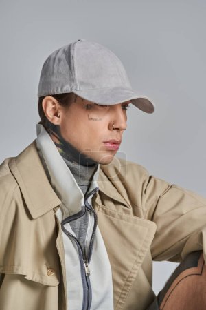 A young, tattooed man stands confidently in a trench coat and hat, exuding an air of mystery and intrigue on a grey background.