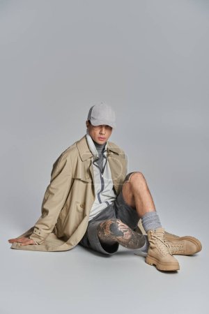 Un jeune homme tatoué dans un trench coat assis par terre les jambes croisées dans un studio sur fond gris.