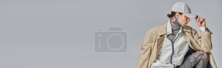 Photo for A young tattooed man exudes mystery and style in a trench coat and hat, standing confidently in a studio against a grey backdrop. - Royalty Free Image