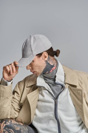 Téléchargez les photos : Un jeune homme tatoué sur son visage s'assoit, vêtu d'un trench coat, dans un studio sur fond gris. - en image libre de droit