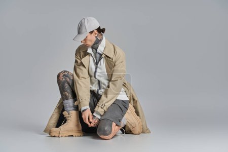 Ein junger Mann mit Tätowierungen sitzt mit Hut und Trenchcoat auf dem Boden vor grauem Studiohintergrund..