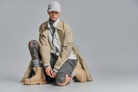 A young, tattooed man sits on the ground, solemnly wrapped in a trench coat against a grey studio backdrop.