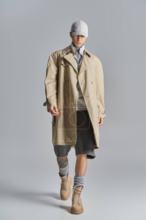 A young, tattooed man showcases an edgy look as he confidently walks down a runway in a trench coat and hat.