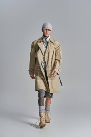 A stylish, tattooed young man in a trench coat poses confidently in a studio against a grey backdrop.