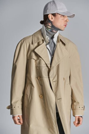 A young, tattooed man exudes mystery and intrigue in a trench coat, donning a stylish hat against a grey studio backdrop.