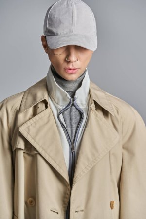 Téléchargez les photos : Un jeune homme tatoué en trench-coat et chapeau se tient en confiance dans un studio sur fond gris. - en image libre de droit