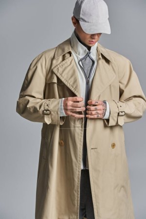 Un jeune homme tatoué respire le mystère et le style dans un trench-coat et un chapeau sur fond gris.