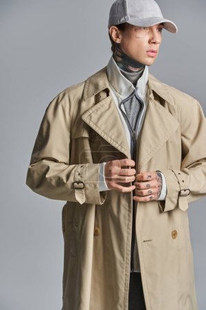 A young, tattooed man exudes an air of mystery as he poses in a trench coat and hat against a grey backdrop in a studio.