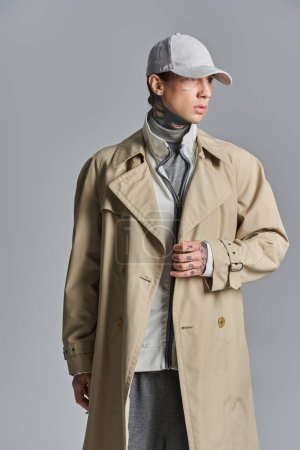 Ein junger, tätowierter Mann trägt einen stylischen Trenchcoat und Hut und verströmt in einem Studio-Ambiente eine geheimnisvolle und urbane Atmosphäre.