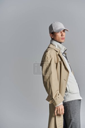 Ein junger, tätowierter Mann mit Hut und Trenchcoat steht selbstbewusst in einem Studio vor grauem Hintergrund..
