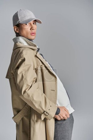 Ein stilvoller Mann mit markantem Hut und elegantem Trenchcoat posiert selbstbewusst in einem Studio vor grauem Hintergrund.