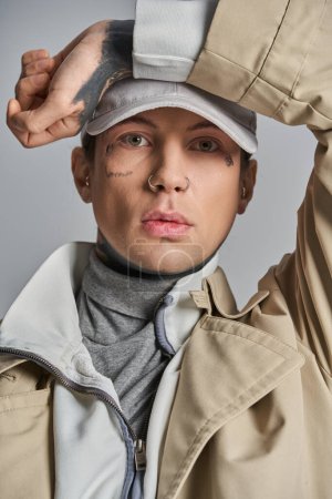 Téléchargez les photos : Un jeune homme tatoué en trench-coat se tient debout avec confiance, portant un chapeau élégant sur la tête dans un décor de studio sur fond gris. - en image libre de droit