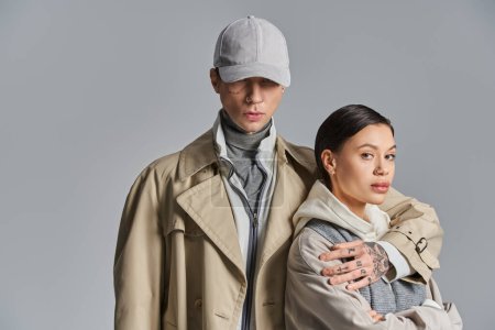 Photo for A young man and woman stand side by side in trench coats, exuding style and grace in a studio against a grey background. - Royalty Free Image