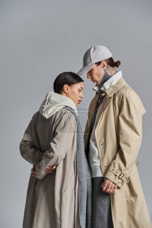 A young stylish couple in trench coats standing together in a studio on a grey background.