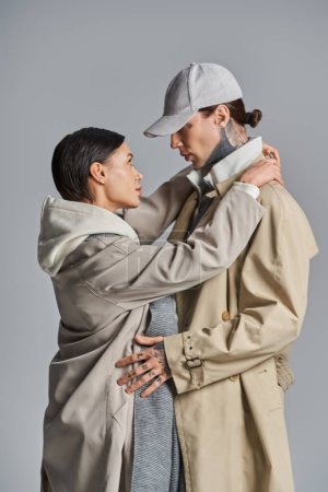 Una pareja joven y elegante, una en una gabardina, la otra en un sombrero, posando en un estudio sobre un fondo gris.