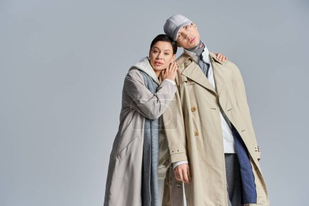 Foto de Una pareja joven y elegante se encuentra lado a lado, exudando elegancia en abrigos de trinchera, en un estudio sobre un fondo gris. - Imagen libre de derechos