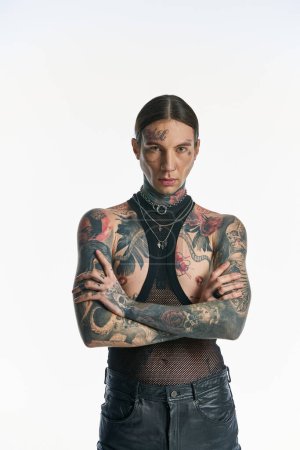Un joven con estilo y tatuajes se levanta con confianza, con los brazos cruzados, en un estudio sobre un fondo gris.
