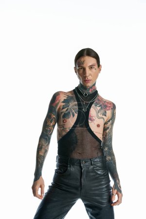 A tattooed man exudes attitude in leather pants, showcasing his bold style against a grey studio backdrop.