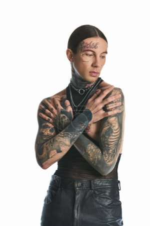 Un jeune homme tatoué ornant ses bras et sa poitrine pose élégamment dans un studio sur fond gris.
