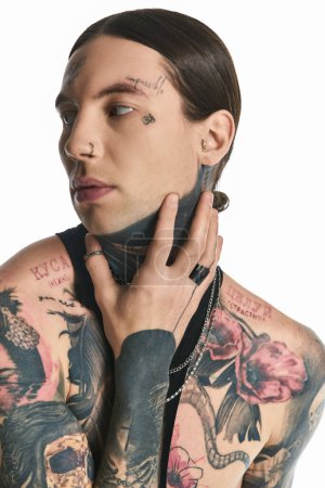 Ein junger, stylischer Mann mit einer Vielzahl von Tätowierungen auf Körper und Hals posiert in einem Studio vor grauem Hintergrund.