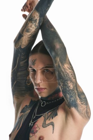 A young stylish man with extensive tattoos and piercings on his arms poses in a studio against a grey background.