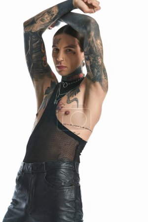 A young man with tattoos on his arm and chest poses in a studio against a grey background.