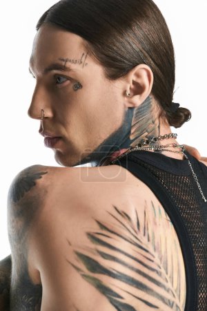 Un jeune homme élégant arborant fièrement un tatouage sur son épaule, dans un décor studio sur fond gris.
