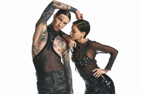 A young, stylish and tattooed man and woman posing together in a studio against a grey background.