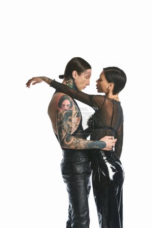 A stylish and tattooed young couple sharing a warm hug in a studio setting against a grey background.
